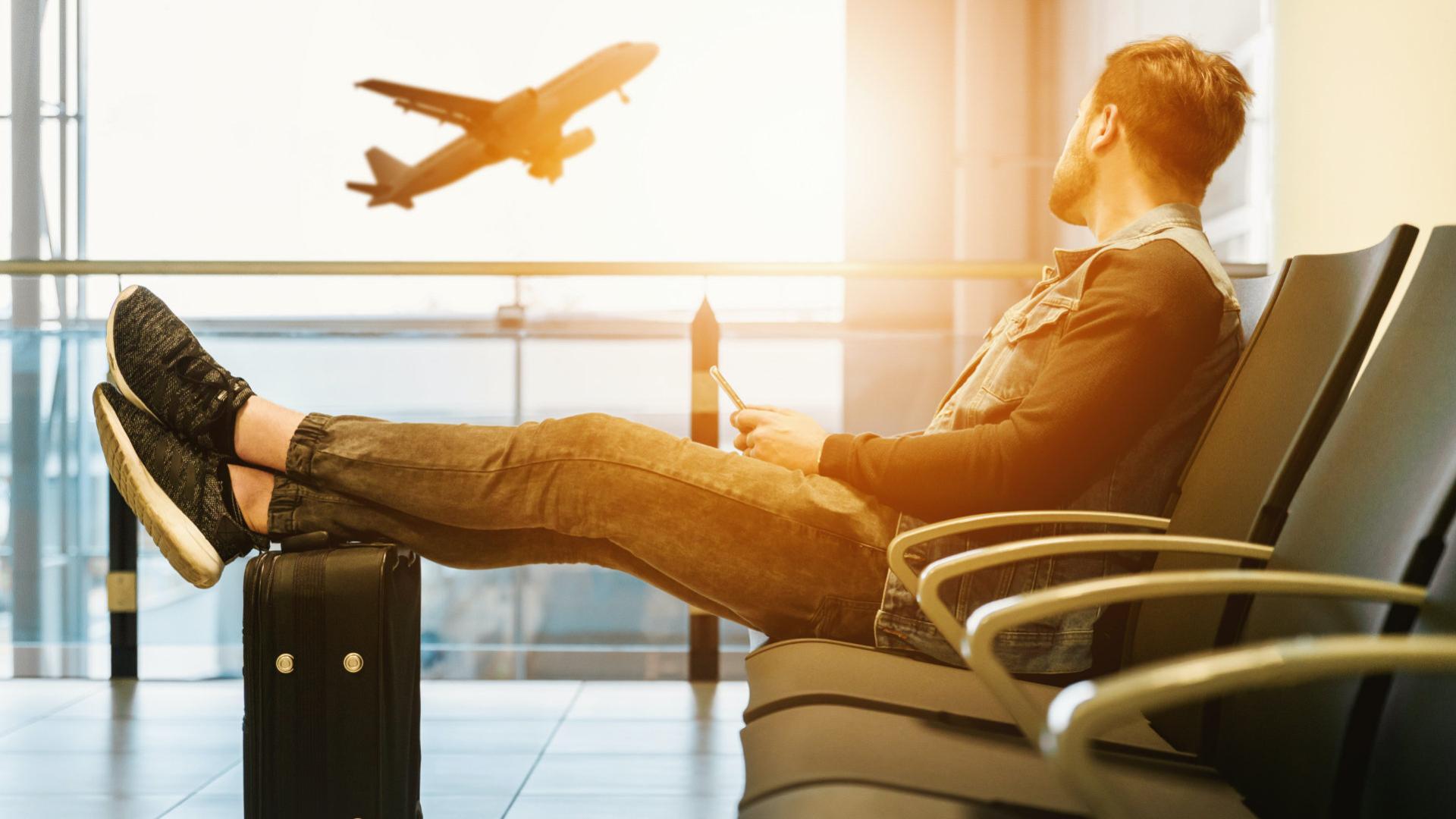 Fotografia de um homem no aeroporto com as pernas sobre a mala, e um avião decolando ao por do sol